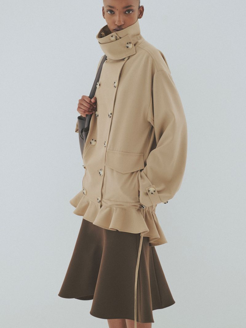 Model wearing a tan double-breasted collared jacket and a brown skirt against a plain light grey background