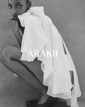 Fashion editorial of a Black woman wearing a white chiffon dress. She is sitting and facing forward in a studio with gray backdrop.
