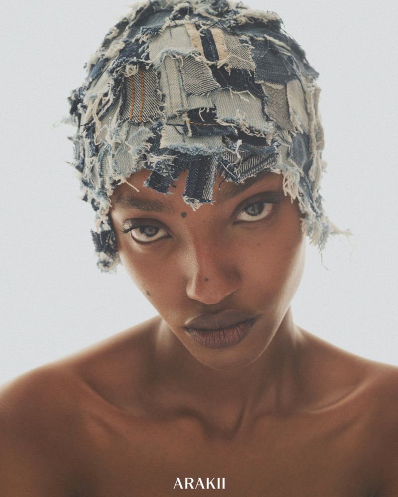 Striking portrait of a woman with tan skin, wearing a patchwork denim hat, looking directly at the camera in a bright studio setting.