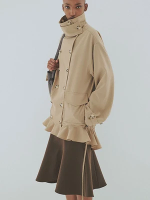 Model wearing a tan double-breasted collared jacket and a brown skirt against a plain light grey background