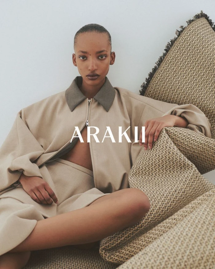 Model wearing tan zip jacket and matching shorts leaning against a textured cushion against a plain white wall