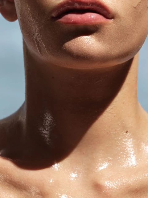 Close-up portrait of a young adult with shiny skin and glossy lips against a light blue background, casting strong shadows.