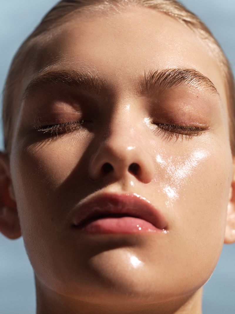 Close-up studio shot of a fair-skinned young woman with closed eyes, shiny skin, and pink lips against a blurred light-blue background.