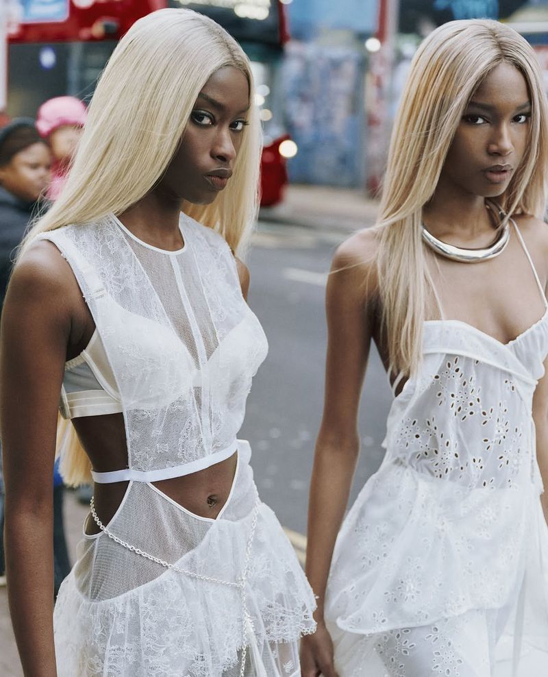 Two women with blonde hair wearing white dresses pose outdoors. Focus on fashion, style, and urban environment. Medium shot, eye-level angle.