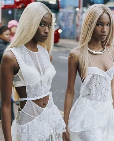 Two women with blonde hair wearing white dresses pose outdoors. Focus on fashion, style, and urban environment. Medium shot, eye-level angle.
