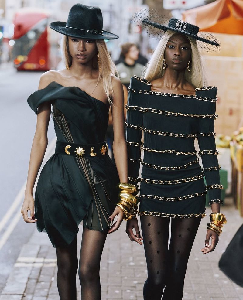 Two fashion models in black dresses and hats pose on a city street with neutral expressions, showcasing couture style and accessories.
