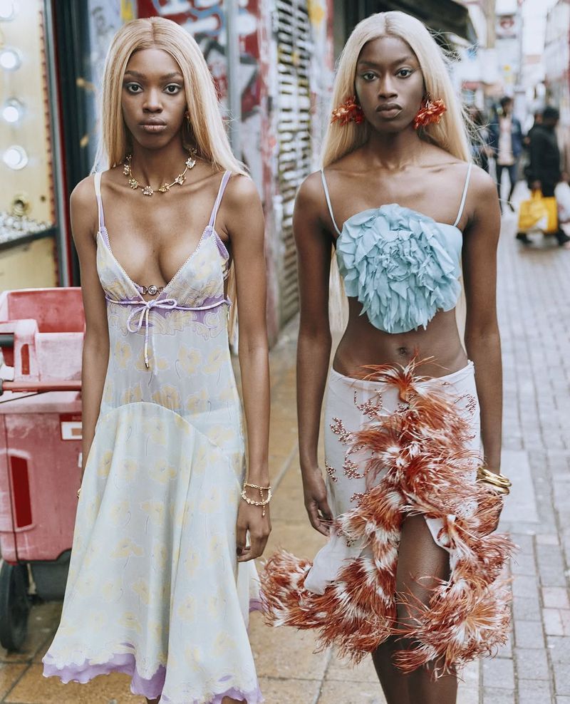 Two women with blonde hair in stylish outfits pose on a city street with a building and graffiti-covered wall in the background.