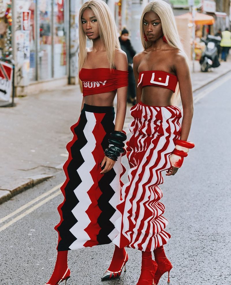 Two fashion models pose outdoors in matching red and white outfits with blonde wigs on a city street with blurred buildings.