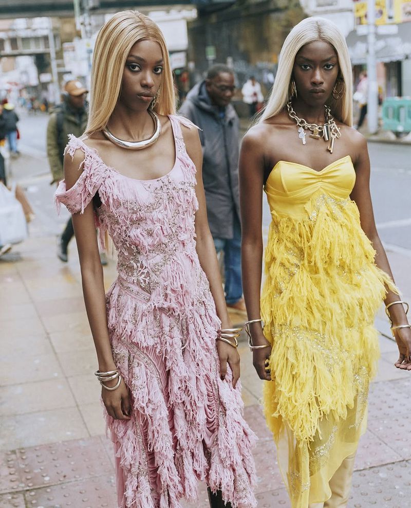Two black female models wearing fringed gowns and blonde wigs pose outdoors in a fashion editorial with pedestrians in background.