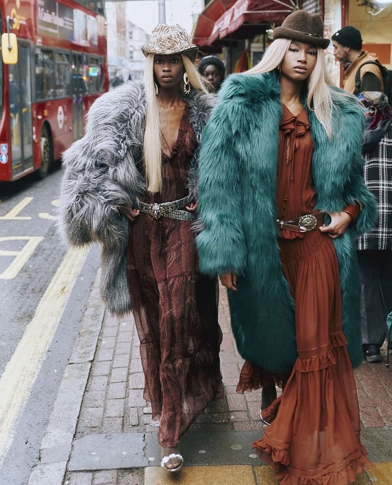 Two women in fur coats, long dresses, and hats walk on a city sidewalk near a red bus and shops, creating a high-fashion street style image.