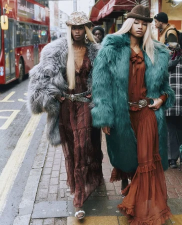 Two women in fur coats, long dresses, and hats walk on a city sidewalk near a red bus and shops, creating a high-fashion street style image.
