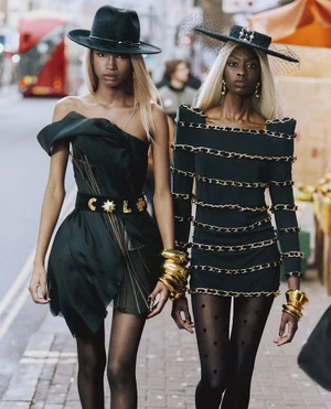 Two models wearing dark designer dresses, wide-brimmed hats, and gold bracelets walking outdoors on a city street.