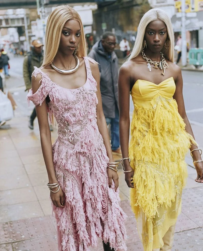 Two models wearing pink and yellow fringed dresses standing on a city street for Dazed Magazine editorial photography by Ib Kamara
