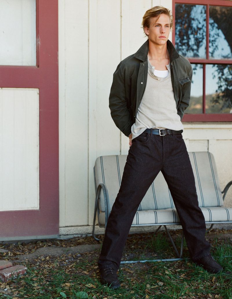 Young adult male fashion model in casual clothes poses outdoors in front of a white wall, wearing a jacket, t-shirt, and dark pants.