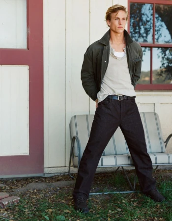 Young adult male fashion model in casual clothes poses outdoors in front of a white wall, wearing a jacket, t-shirt, and dark pants.