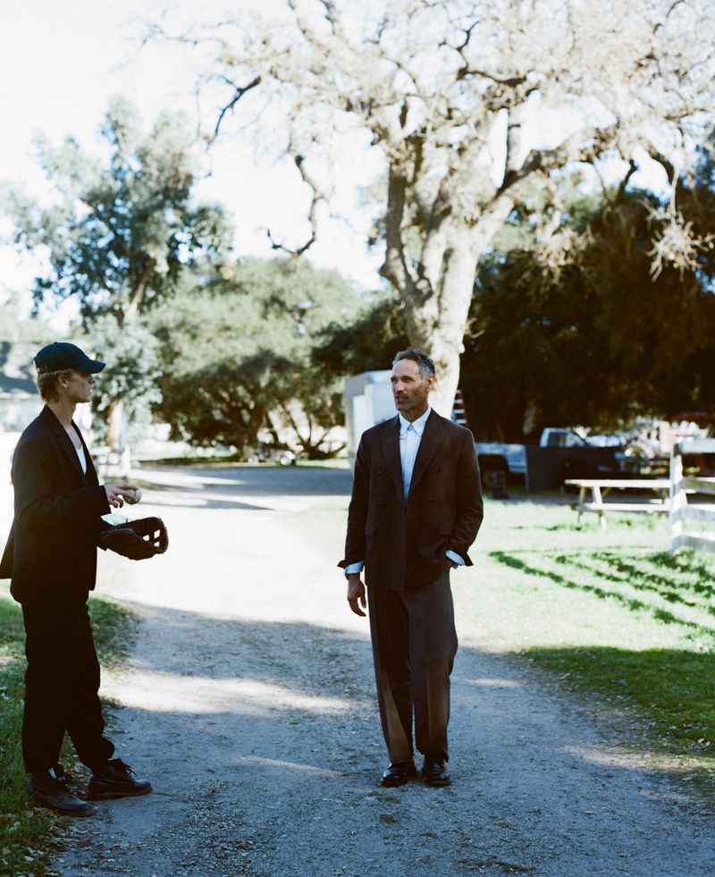 Two men stand on a dirt road in an outdoor setting, one wearing a suit and the other a baseball cap, surrounded by trees and greenery.