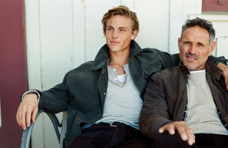 A young adult man and an older man sit together outdoors, wearing casual jackets and shirts in a relaxed pose, creating a natural portrait.