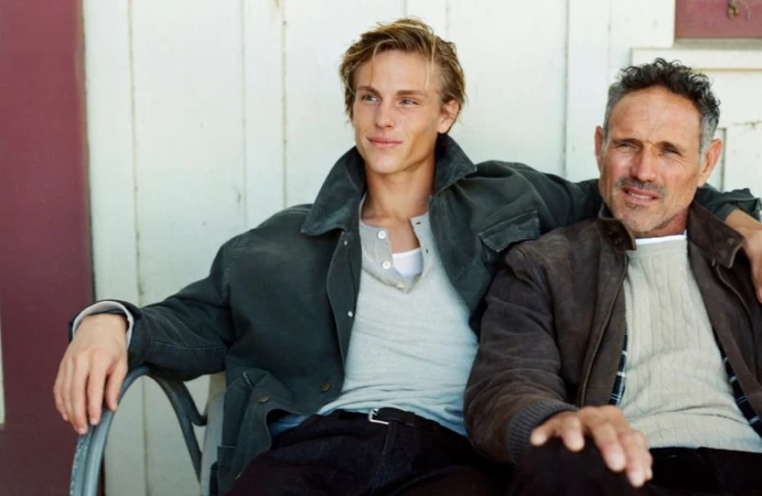 A young adult man and an older man sit together outdoors, wearing casual jackets and shirts in a relaxed pose, creating a natural portrait.