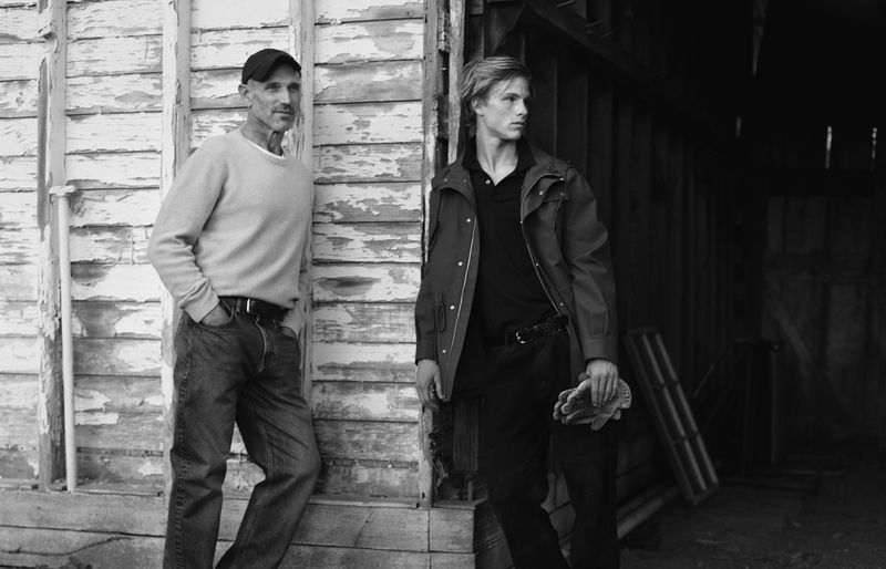 Black and white shot of two men in casual clothing standing in front of a weathered wooden structure with a dark interior space.
