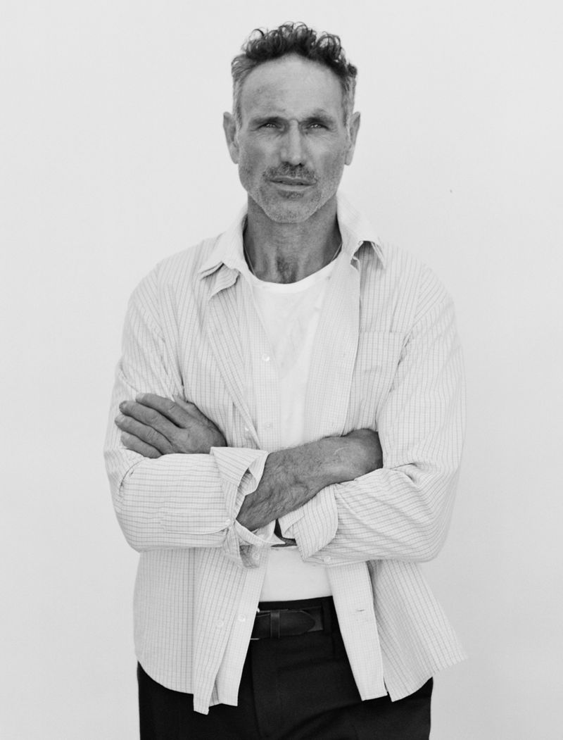 Black and white portrait of a middle-aged man with short hair, wearing an open striped shirt over a t-shirt, standing with arms crossed.