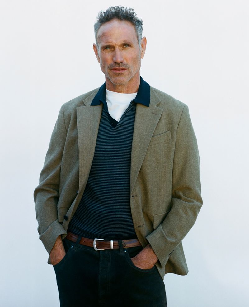 Man in blazer, striped shirt, and dark jeans poses in front of a white wall. Portrait captures his gaze, casual attire, and confident stance.