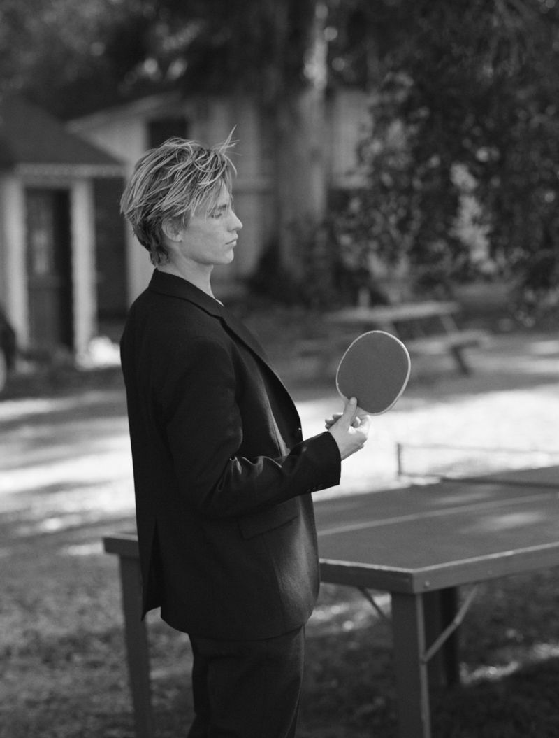Monochrome portrait of a young adult holding a ping pong paddle next to a table outdoors, set against a backdrop of blurred trees and a building.
