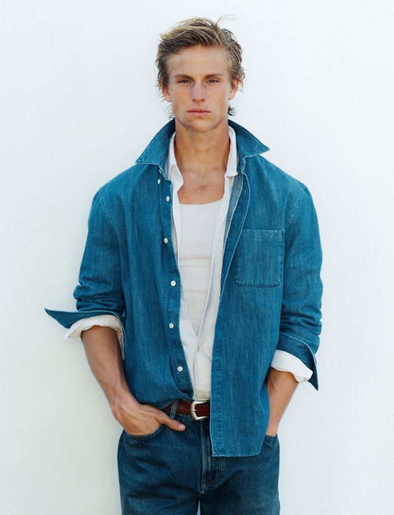 Portrait of a young adult male in denim shirt and jeans against a white wall, posing with hands in pockets in an indoor setting.