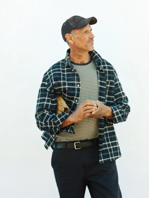 Man in black cap and plaid shirt stands against white background, holding baseball glove, looking off to the side in natural daylight.