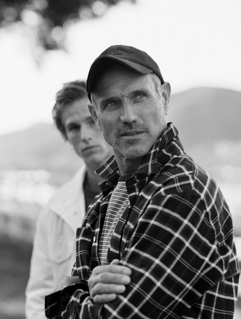 Black and white portrait of a man wearing a plaid shirt and cap with another man in the background