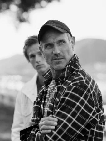 Black and white portrait of a man wearing a plaid shirt and cap with another man in the background
