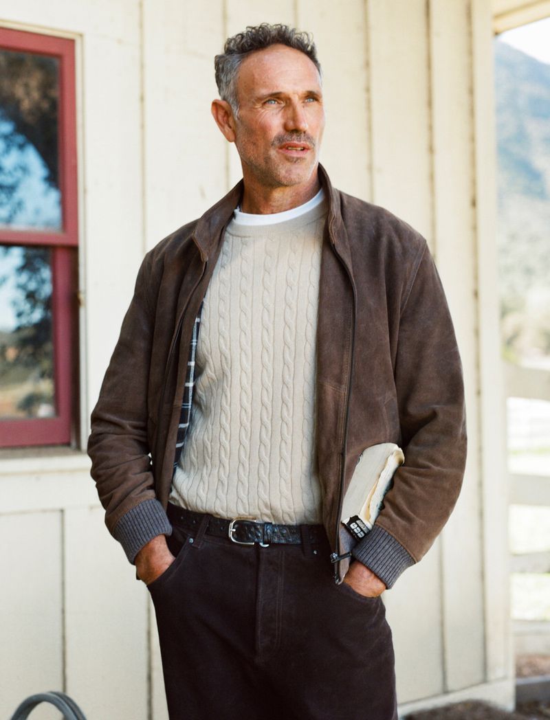 An adult man in casual attire standing outdoors. He wears a suede jacket, cable knit sweater, and jeans, posing against a building and landscape.