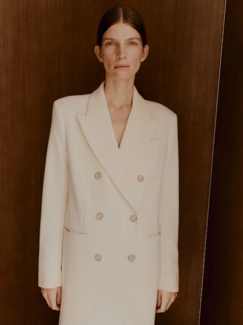 Portrait of a poised adult female in a cream-colored blazer-dress, styled hair, against a neutral brown background, in a studio setting.
