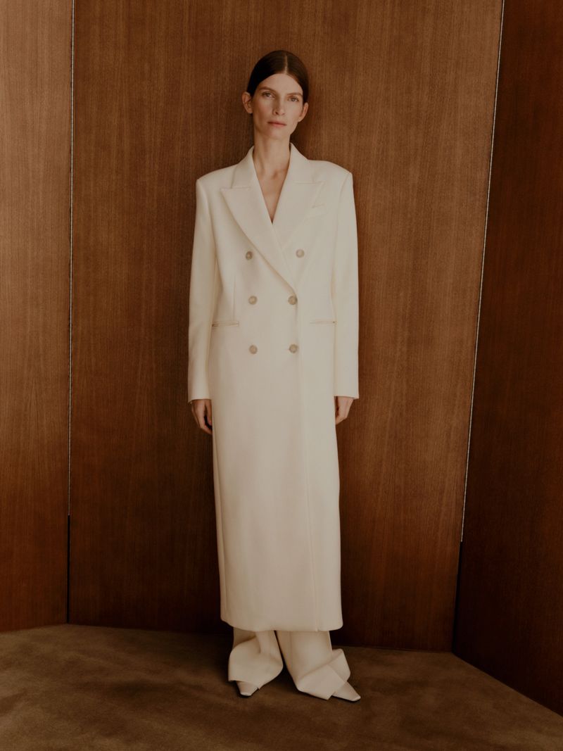 Model standing against wood paneled folding screens wearing a long white double breasted coat and trousers.