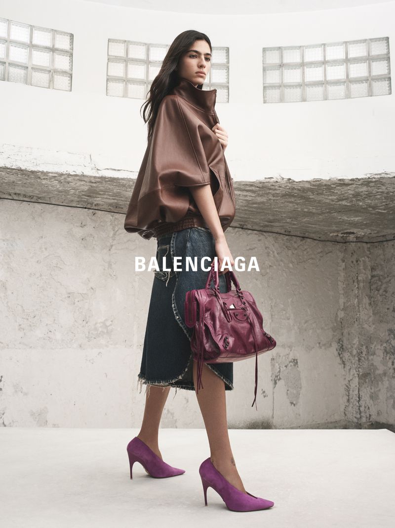 Fashion shot of a woman in a brown leather jacket, denim skirt, and purple heels, holding a purple bag in an indoor setting.