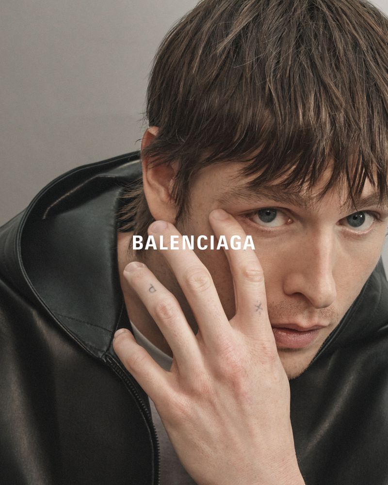Close-up of a male model in a black leather jacket posing in a studio with the Balenciaga logo across his eye and finger tattoos.