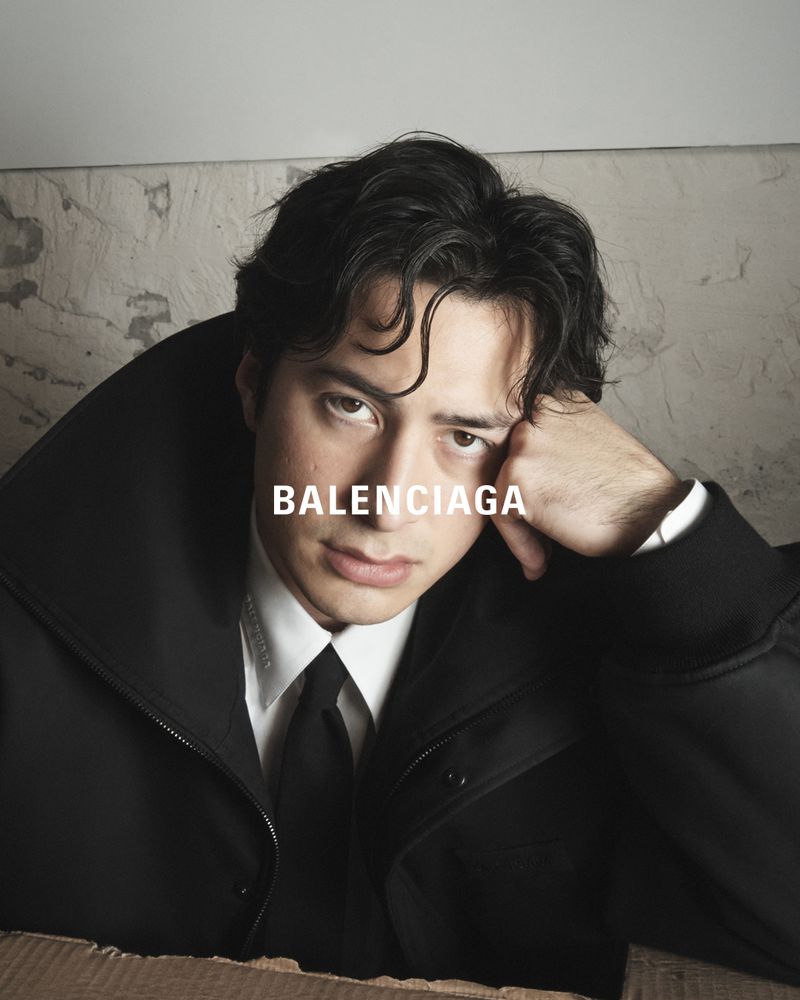 Portrait of a young adult male with dark hair wearing a black suit and tie, resting his face on his hand against a muted background.