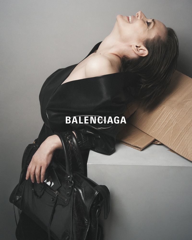 Elegant female model poses with a Balenciaga purse and black robe in a grey studio setting, showcasing fashion and style with confidence.