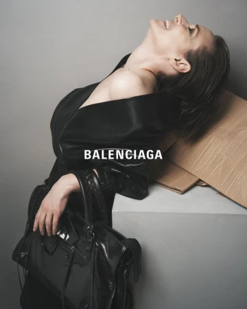 Elegant female model poses with a Balenciaga purse and black robe in a grey studio setting, showcasing fashion and style with confidence.