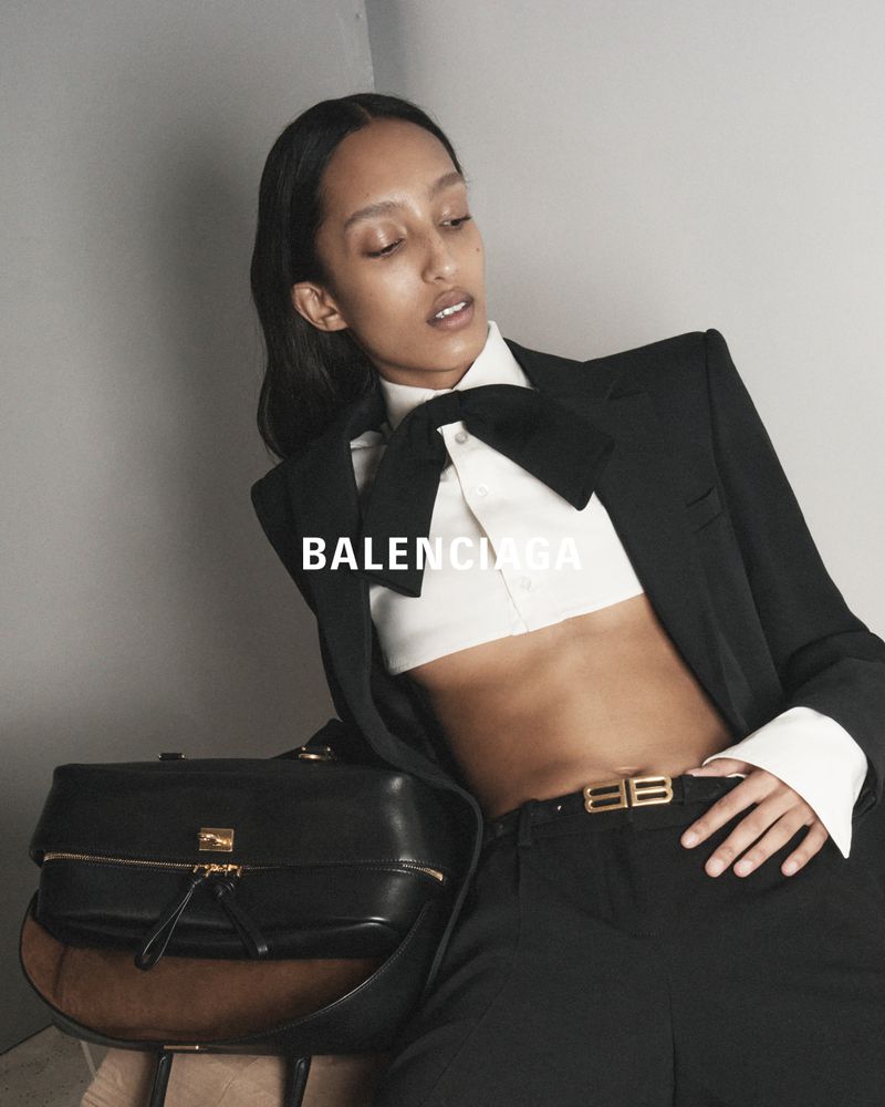 A woman poses indoors with a black blazer, white crop top, black pants and bag, with a black bow tie and Balenciaga belt.