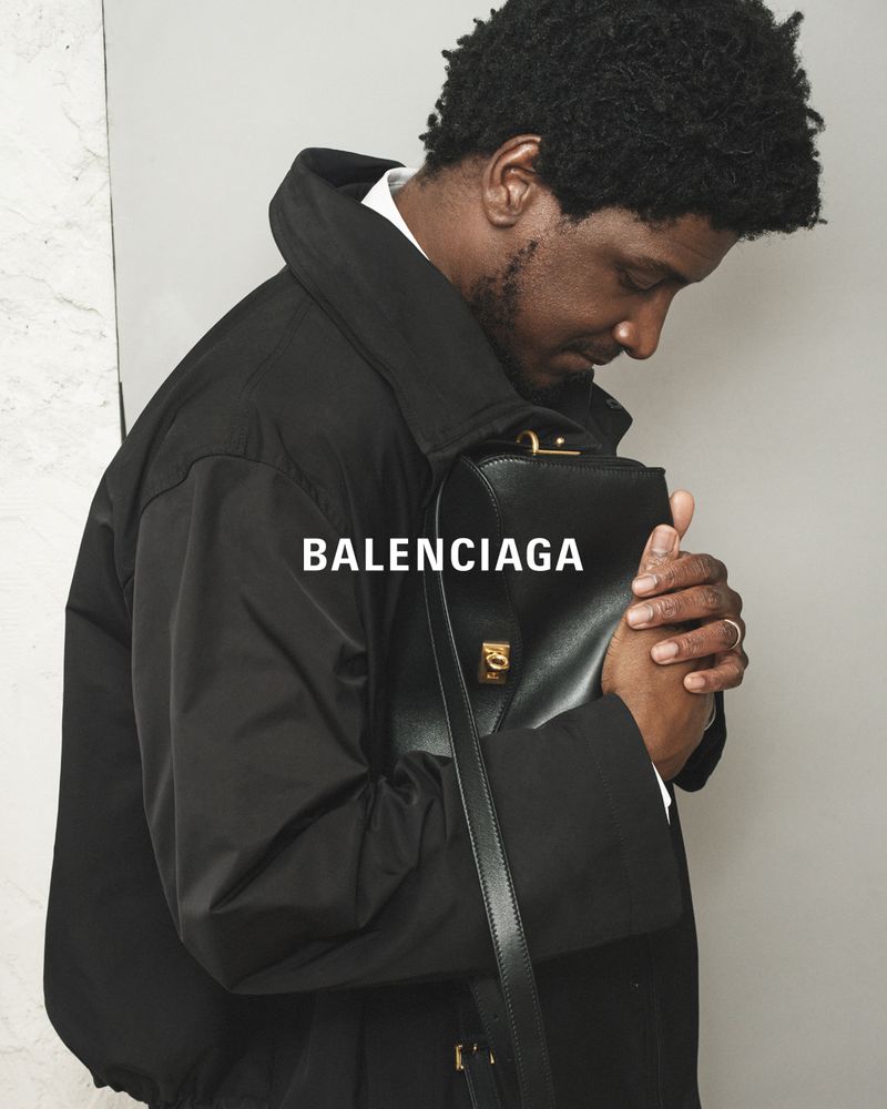 Man in black coat holds Balenciaga bag, showcasing fashion and style against a grey background in a medium shot, suitable for editorials.