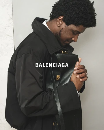 Man in black coat holds Balenciaga bag, showcasing fashion and style against a grey background in a medium shot, suitable for editorials.