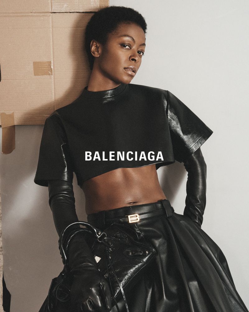 Fashion shot of a Black woman wearing a cropped black top, leather skirt, and long gloves, posing against a cardboard box background.