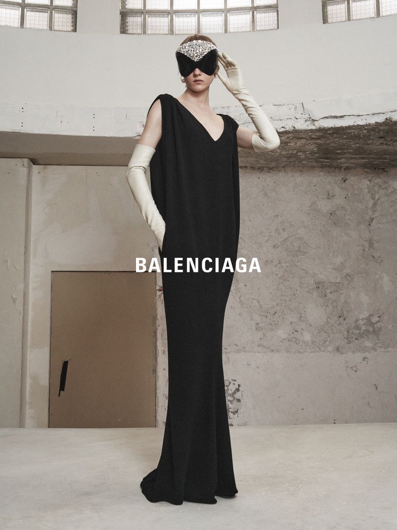 Elegant woman in a black gown and jeweled mask, posing in an editorial fashion shot against a textured concrete background.