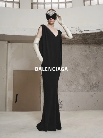 Elegant woman in a black gown and jeweled mask, posing in an editorial fashion shot against a textured concrete background.