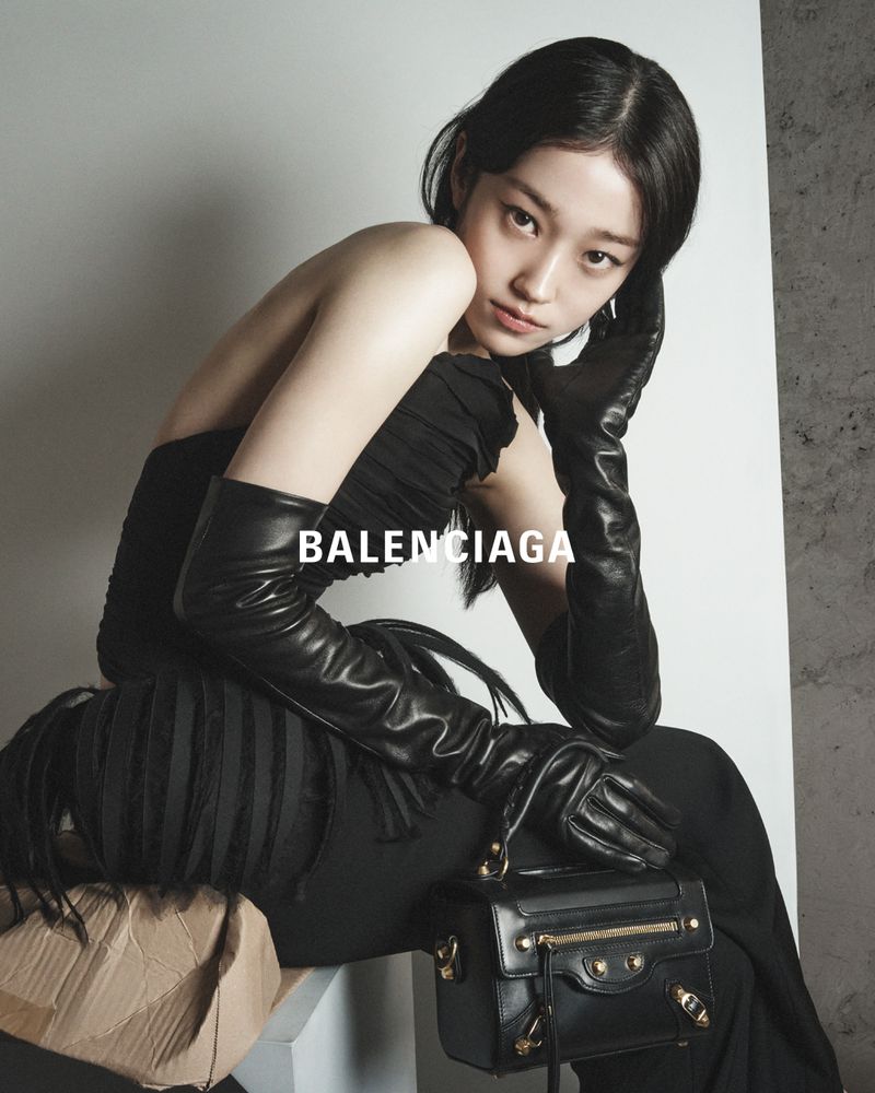 A stylish Asian woman poses in a black dress, gloves, and handbag by Balenciaga, creating a fashion-forward image with neutral studio backdrop.