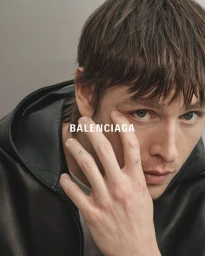 Close-up portrait of a young man wearing a black leather jacket against a grey background with Balenciaga text