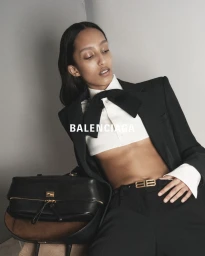 Model in black suit jacket and cropped white shirt with bow tie resting hand on black leather bag