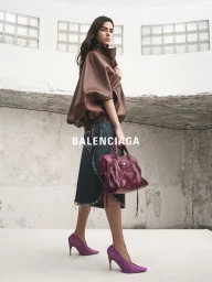 Model in brown leather jacket and denim skirt holding a magenta handbag against a concrete architectural background