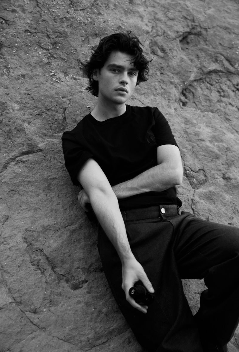 Monochrome portrait of a young adult male with dark hair in a black t-shirt and dark pants leaning against a rock formation.