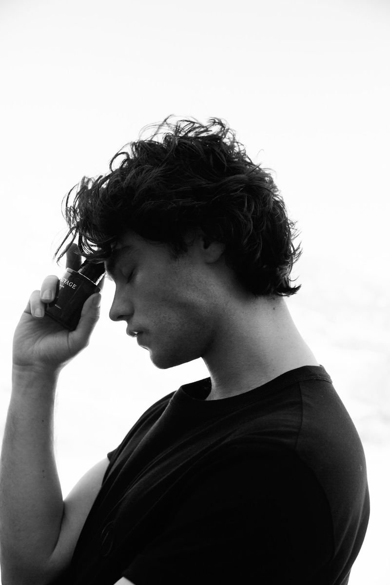 Black and white studio portrait of a young adult male holding a black bottle against his head, against a bright white background.